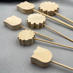 May include: A collection of wooden cocktail picks in various shapes, including flower and cat silhouettes. The picks are made of light-colored wood and are attached to thin bamboo skewers. The items are arranged on a gray surface.