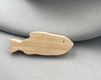 May include: A wooden fish-shaped ornament with a natural wood finish. The fish has a small, round eye and a tail fin. The ornament is lying on a gray fabric surface.