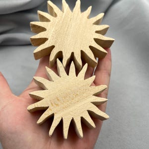 Może przedstawiać: Dwa niewykończone drewniane kształty słońca z ostrymi promieniami. Drewno ma jasny, naturalny kolor i widoczne usłojenie. Słońca są trzymane w dłoni, co sugeruje projekt rzemieślniczy lub DIY.