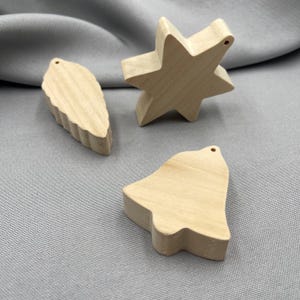 May include: Three unfinished wooden ornaments: a leaf, a star, and a bell. Each ornament has a small hole at the top for hanging. The leaf has a textured edge, while the star is multi-pointed. The bell has a rounded bottom.