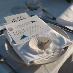 Op de afbeelding: Een tafelsetting voor een bruiloft met een wit bord, servet en bestek. Een menukaart met de tekst "ALL I SEA IS LOVE AND GOOD FOOD" en "WAVES OF WELCOME" ligt op het bord. Een zeeschelp met de naam "Ardeen" ligt op het servet.