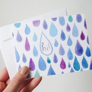 May include: A white card with a blue, purple, and green watercolor raindrop pattern. The word "hi!" is written in black ink inside one of the raindrops.