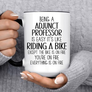 May include: White ceramic mug with a black handle and rim, featuring the text "BEING A ADJUNCT PROFESSOR IS EASY IT'S LIKE RIDING A BIKE EXCEPT THE BIKE IS ON FIRE YOU'RE ON FIRE EVERYTHING IS ON FIRE".