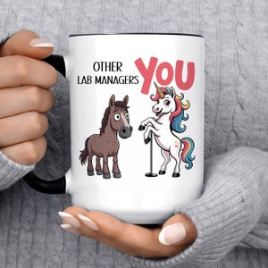 May include: White ceramic mug with a black handle and rim. The mug features a cartoon illustration of a brown horse and a unicorn with a rainbow mane and tail. Text on the mug reads "OTHER LAB MANAGERS" and "You" in pink.