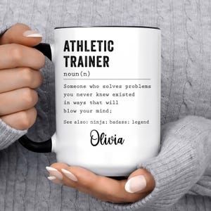 May include: White ceramic mug with black text that reads "ATHLETIC TRAINER noun (n) Someone who solves problems you never knew existed in ways that will blow your mind; See also: ninja; badass; legend Olivia"