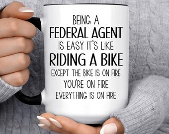 May include: White ceramic mug with a black rim and handle. The mug has black text that reads "Being a Federal Agent is easy it's like riding a bike except the bike is on fire you're on fire everything is on fire".