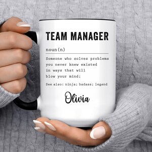May include: A white ceramic mug with a black handle and rim, featuring the black printed text: TEAM MANAGER noun(n) Someone who solves problems you never knew existed in ways that will blow your mind; See also: ninja; badass; legend. The name Olivia is printed below.