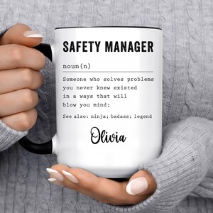 May include: White ceramic mug with black text that reads "SAFETY MANAGER noun (n) Someone who solves problems you never knew existed in a ways that will blow you mind; See also: ninja; badass; legend Olivia".