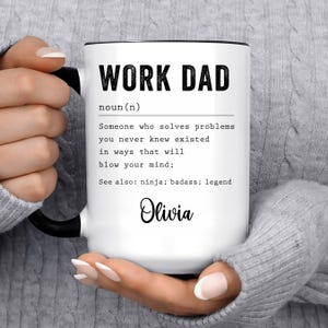 May include: White ceramic mug with a black rim and handle. The mug features a black and white graphic with the text "WORK DAD" and a definition of the term. The name "Olivia" is written in cursive at the bottom of the mug.