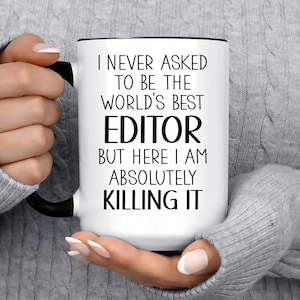 May include: White ceramic mug with a black handle and rim, featuring the text "I NEVER ASKED TO BE THE WORLD'S BEST EDITOR BUT HERE I AM ABSOLUTELY KILLING IT" in black font. The mug is held by a person wearing a gray sweater.