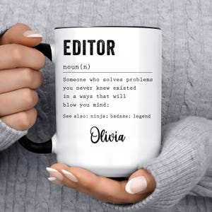 May include: White ceramic mug with a black rim and handle. The mug features a definition of the word "editor" with the name "Olivia" at the bottom. The text reads: "EDITOR noun (n) Someone who solves problems you never knew existed in a ways that will blow you mind; See also: ninja; badass; legend Olivia"