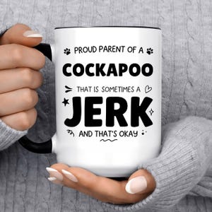 May include: A white ceramic mug with a black handle and rim, featuring black text that reads: PROUD PARENT OF A COCKAPOO THAT IS SOMETIMES A JERK AND THAT'S OKAY, decorated with small paw prints, a heart, a star, and various small doodle accents.