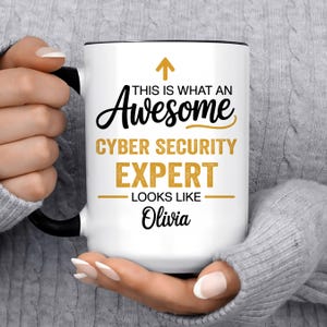 May include: A white ceramic mug with a black handle and rim, featuring the black and gold text: THIS IS WHAT AN Awesome CYBER SECURITY EXPERT LOOKS LIKE Olivia, with an upward-pointing gold arrow above the text and horizontal gold lines.