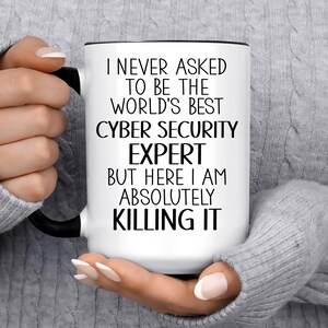 May include: A white ceramic mug with a black handle and rim, featuring the black printed text: I NEVER ASKED TO BE THE WORLD'S BEST CYBER SECURITY EXPERT BUT HERE I AM ABSOLUTELY KILLING IT. The mug has a smooth finish and a cylindrical shape.