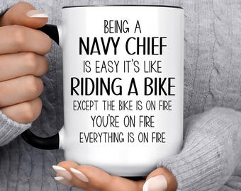 May include: White ceramic mug with a black handle and rim. The mug features the text "BEING A NAVY CHIEF IS EASY IT'S LIKE RIDING A BIKE EXCEPT THE BIKE IS ON FIRE YOU'RE ON FIRE EVERYTHING IS ON FIRE" in black.