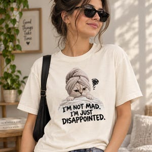 May include: A cream-colored short-sleeve t-shirt featuring a graphic of a cat wearing a gray towel on its head, resting on a white surface. Below the cat, the text reads, "I'M NOT MAD. I'M JUST DISAPPOINTED." with a small scribble icon.