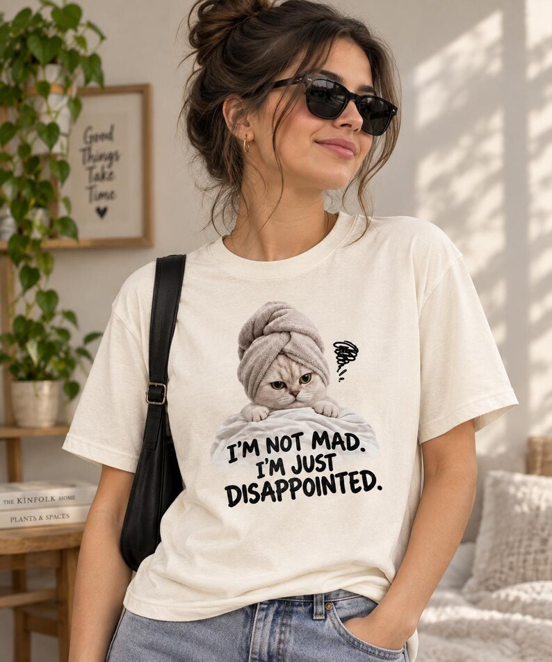 May include: A cream-colored short-sleeve t-shirt featuring a graphic of a cat wearing a gray towel on its head, resting on a white surface. Below the cat, the text reads, "I'M NOT MAD. I'M JUST DISAPPOINTED." with a small scribble icon.