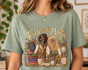 May include: A sage green t-shirt featuring a graphic print with the text "Teach with Magic". The illustration depicts two individuals from behind surrounded by flowers, books, a globe, a chalkboard, and mugs. The books are labelled Literature, Math, Science, Art, and History.