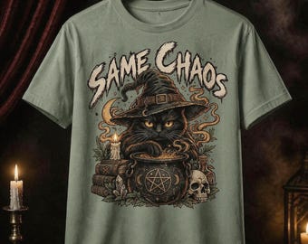 May include: A sage green t-shirt featuring a graphic print of a black cat wearing a witch hat, stirring a cauldron with a pentagram symbol. The text "SAME CHAOS" is printed above the cat. The design includes a skull, books, and candles.