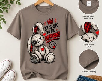 May include: A brownish-gray short-sleeve t-shirt featuring a graphic of a stitched rabbit with red plaid ears, a red heart, and a red crown. The text reads IT'S OK TO BE DIFFERENT above a circular smiley face with X eyes and a tongue sticking out.