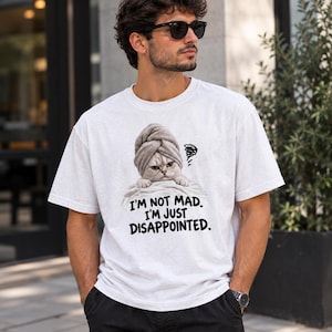 May include: A white short-sleeved t-shirt featuring a graphic of a cat wearing a grey towel on its head. The cat has a grumpy expression and is positioned above the black text that reads: I'M NOT MAD. I'M JUST DISAPPOINTED. The shirt has a crew neckline.