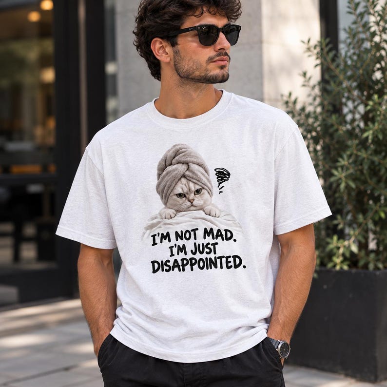 May include: A white short-sleeved t-shirt featuring a graphic of a cat wearing a grey towel on its head. The cat has a grumpy expression and is positioned above the black text that reads: I'M NOT MAD. I'M JUST DISAPPOINTED. The shirt has a crew neckline.
