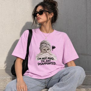 May include: A light pink short-sleeved t-shirt featuring a graphic of a cat wearing a towel on its head. The text below the cat reads, I'M NOT MAD. I'M JUST DISAPPOINTED. The shirt is paired with a black shoulder bag and blue denim jeans.