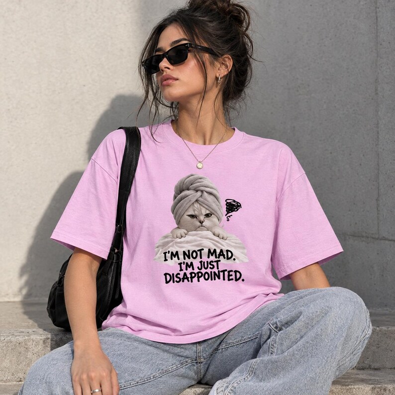 May include: A light pink short-sleeved t-shirt featuring a graphic of a cat wearing a towel on its head. The text below the cat reads, I'M NOT MAD. I'M JUST DISAPPOINTED. The shirt is paired with a black shoulder bag and blue denim jeans.