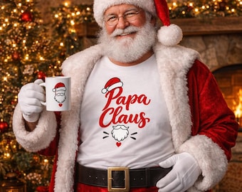 May include: A white t-shirt featuring the red text Papa Claus, a graphic of a Santa hat, a white beard, and a small red heart. The shirt is worn by a person dressed in a traditional red and white Santa suit with a matching hat and white gloves.