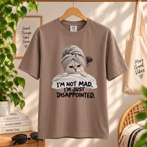 May include: A taupe short-sleeve t-shirt featuring a graphic of a cat wearing a gray towel on its head, resting on a white towel. The text below the cat reads, "I'M NOT MAD. I'M JUST DISAPPOINTED." The shirt has a crew neckline and a standard fit.