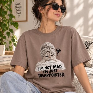 May include: A brown short-sleeved t-shirt featuring a graphic of a cat wearing a towel on its head, resting on a white pillow. The text below the cat reads, "I'M NOT MAD. I'M JUST DISAPPOINTED." with a small scribble icon above the cat's head.