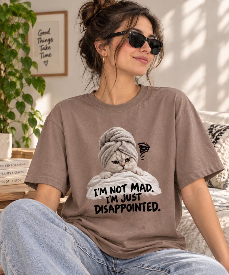 May include: A brown short-sleeved t-shirt featuring a graphic of a cat wearing a towel on its head, resting on a white pillow. The text below the cat reads, "I'M NOT MAD. I'M JUST DISAPPOINTED." with a small scribble icon above the cat's head.
