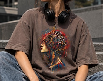 May include: A brown short-sleeved t-shirt featuring a graphic print of a side profile of a face surrounded by abstract, colourful paint splatters in red, yellow, and blue. The design includes swirling lines and drips over the head area.