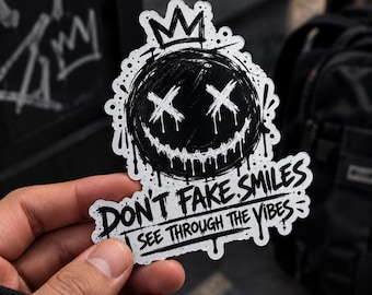 May include: A die-cut sticker featuring a black circular face with a dripping paint effect, X-shaped eyes, and a wide, jagged grin. A crown sits atop the head. Below the face, the text reads: DON'T FAKE SMILES SEE THROUGH THE VIBES.