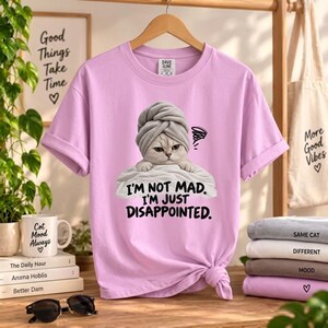 May include: A light purple t-shirt featuring a graphic of a cat wearing a gray towel on its head, resting on a white pillow. The text below the cat reads, "I'M NOT MAD. I'M JUST DISAPPOINTED." The shirt has short sleeves and a crew neckline.