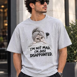 May include: A light gray short-sleeved t-shirt featuring a graphic of a cat wearing a towel on its head, peeking over a blanket. The text below the cat reads, I'M NOT MAD. I'M JUST DISAPPOINTED. The shirt has a crew neckline and a relaxed fit.