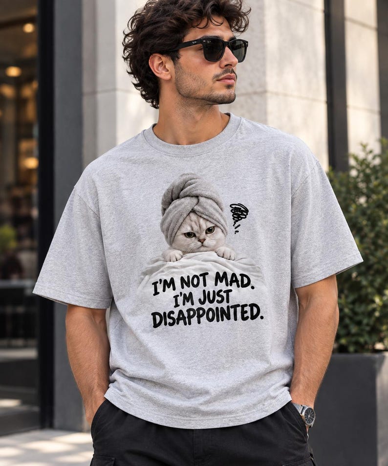 May include: A light gray short-sleeved t-shirt featuring a graphic of a cat wearing a towel on its head, peeking over a blanket. The text below the cat reads, I'M NOT MAD. I'M JUST DISAPPOINTED. The shirt has a crew neckline and a relaxed fit.