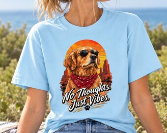 May include: A light blue t-shirt featuring a graphic of a golden retriever wearing black sunglasses and a red patterned bandana. The background shows a desert landscape with a sunset. The text reads No Thoughts. Just Vibes. with a small paw print.