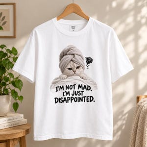 May include: A white short-sleeved t-shirt featuring a graphic of a cat wearing a grey towel on its head. The cat has a grumpy expression and is wrapped in a white towel. Below the cat, the text reads: I'M NOT MAD. I'M JUST DISAPPOINTED.