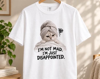 May include: A white short-sleeved t-shirt featuring a graphic of a cat wearing a grey towel on its head. The cat has a grumpy expression and is wrapped in a white towel. Below the cat, the text reads: I'M NOT MAD. I'M JUST DISAPPOINTED.