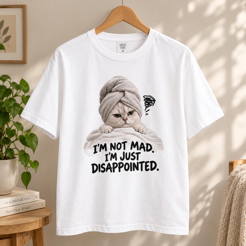 May include: A white short-sleeved t-shirt featuring a graphic of a cat wearing a grey towel on its head. The cat has a grumpy expression and is wrapped in a white towel. Below the cat, the text reads: I'M NOT MAD. I'M JUST DISAPPOINTED.
