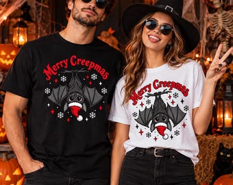 May include: A black t-shirt and a white t-shirt, each featuring a graphic of a bat wearing a red and white Santa hat, hanging from a branch. The text Merry Creepmas is printed in red above the bat, surrounded by white snowflakes and red stars.