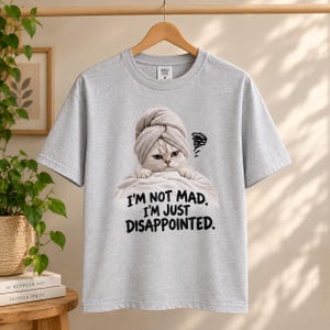 May include: A light gray short-sleeve t-shirt featuring a graphic of a cat wearing a towel on its head, resting on a white cloth. Below the cat, the black text reads, I'M NOT MAD. I'M JUST DISAPPOINTED. The shirt has a crew neckline and a standard fit.