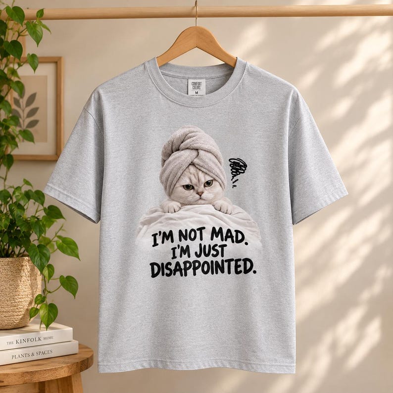May include: A light gray short-sleeve t-shirt featuring a graphic of a cat wearing a towel on its head, resting on a white cloth. Below the cat, the black text reads, I'M NOT MAD. I'M JUST DISAPPOINTED. The shirt has a crew neckline and a standard fit.