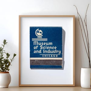 May include: Framed print of a vintage matchbook from the Museum of Science and Industry in Chicago. The matchbook has a dark blue cover with white text and a globe logo. The frame is a light brown color.