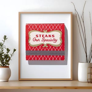 May include: A framed square print featuring a red background with a white diamond lattice pattern. Centered is a cream-colored decorative label with the red text "STEAKS Our Specialty". A horizontal dark gray strike strip sits below the label.