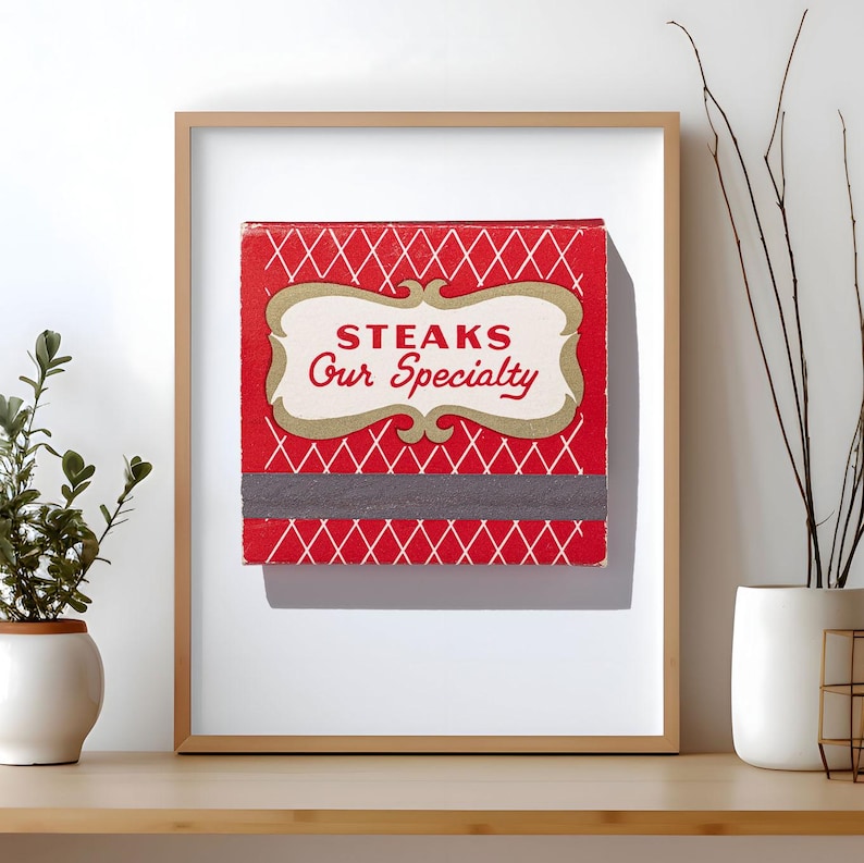 May include: A framed square print featuring a red background with a white diamond lattice pattern. Centered is a cream-colored decorative label with the red text "STEAKS Our Specialty". A horizontal dark gray strike strip sits below the label.