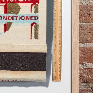 May include: A vintage matchbook with a red and white sign that says "Vision Conditioned". The matchbook is made of paper and has a black match striker on the back. The matchbook is framed and has a ruler next to it.
