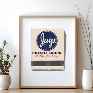 May include: Framed vintage Jays Potato Chips matchbox. The matchbox features a cream-colored background with a blue circular logo that says "Jays". Below the logo, it reads "POTATO CHIPS" and "A Pip of a Chip" in red. The matchbox is framed in a light brown wooden frame.