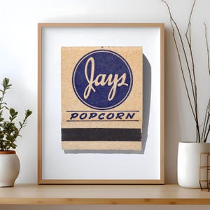 May include: A framed print featuring a vintage-style matchbook cover with a tan background. The design includes a dark blue circle containing the word "jays" in script, with "POPCORN" printed below in blue, and a black strike strip at the bottom.